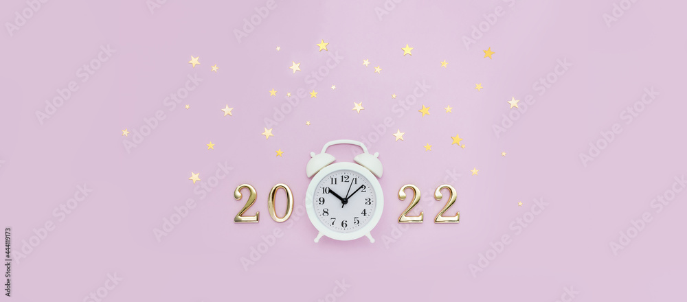 Numbers 2022 and white alarm clock are decorated with star confetti on pink background. Minimal New Year concept. Top view, banner Numbers 2022 and white alarm clock are decorated with star confetti on pink background. Minimal New Year concept. Top view, banner