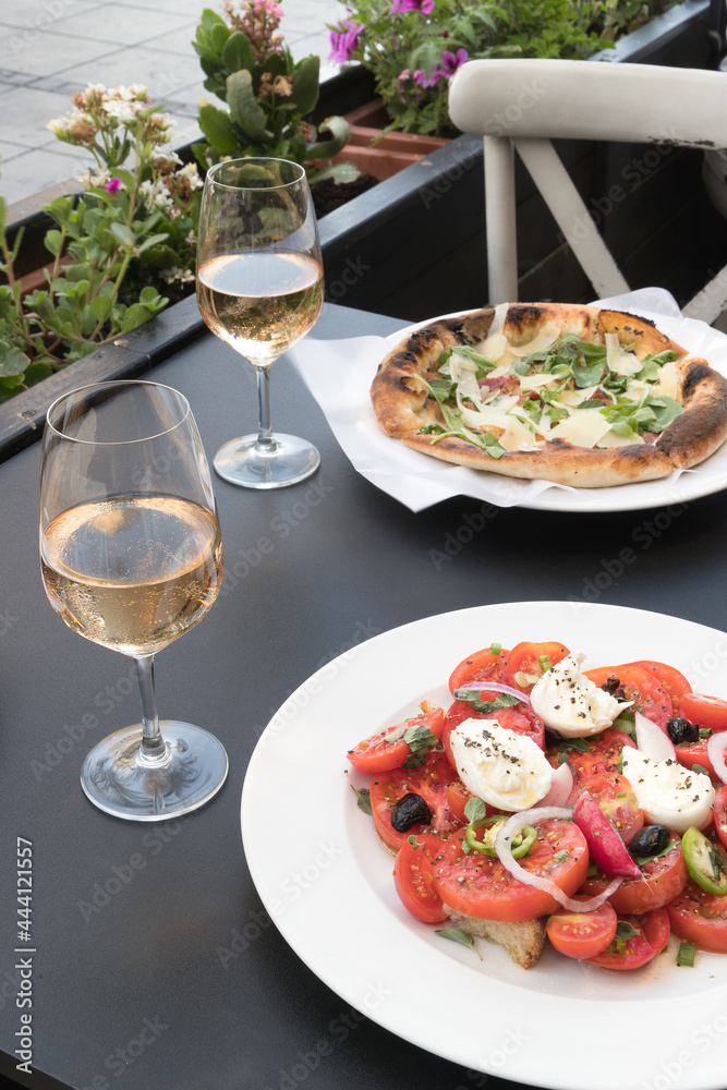Italian caprese salad with tomatoes, mozzarella, oregano, thassos, onions, chili and olive oil. pizza with roccula and parmesan bacon. two glasses of wine rose in a restaurant on the street