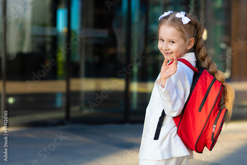 little cute girl in uniform dress, go to first grade, waving hand. happy laughing child back to school. kid with red backpack, schoolbag on campus. copy space, text