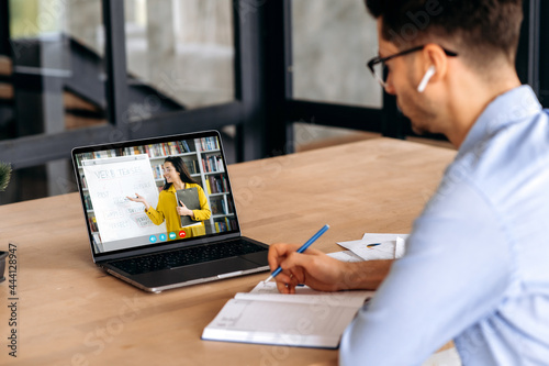 E-learning concept. Clever caucasian male student gains knowledge watching lesson online and studying distantly. Guy takes notes during webinar, listening lecture from female teacher on video call