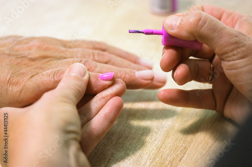 A manicurist applies pink gel shellac nail polish with a brush on a client s nails in a beauty salon. Spa treatments. Women s hands and fingers at the wooden table up close. Female nail care process.