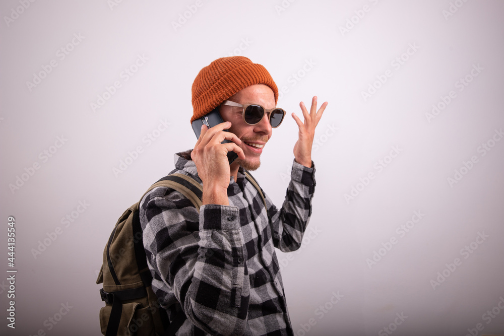 Stylish guy makes a phone call, he wears a plaid shirt, sunglasses and a cap, hipster with backpack, photo taken in the studio with white background. Stylish guy makes a phone call, he wears a plaid shirt, sunglasses and a cap, hipster with backpack, photo taken in the studio with white background.