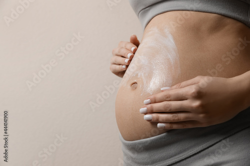 Fotografie Pregnant woman applying cosmetic product on belly against beige background, closeup