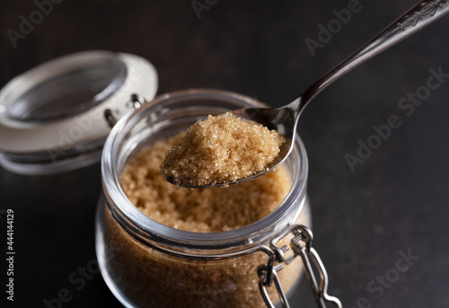 Brown sugar and a spoon in a glass container set against a black stone background.