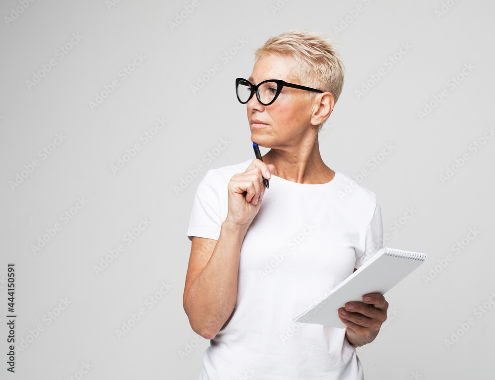 Photo of happy lovely smiling mature woman author in glasses writing in notebook isolated on grey color background