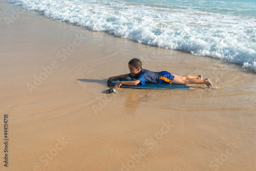 A boy lay down on body board while the wave move to the beach..Paradise beach blue sea, and clear sand landscape. .waves crashing on the beach background..smiling face of happy boy relax concept. © Narong Niemhom