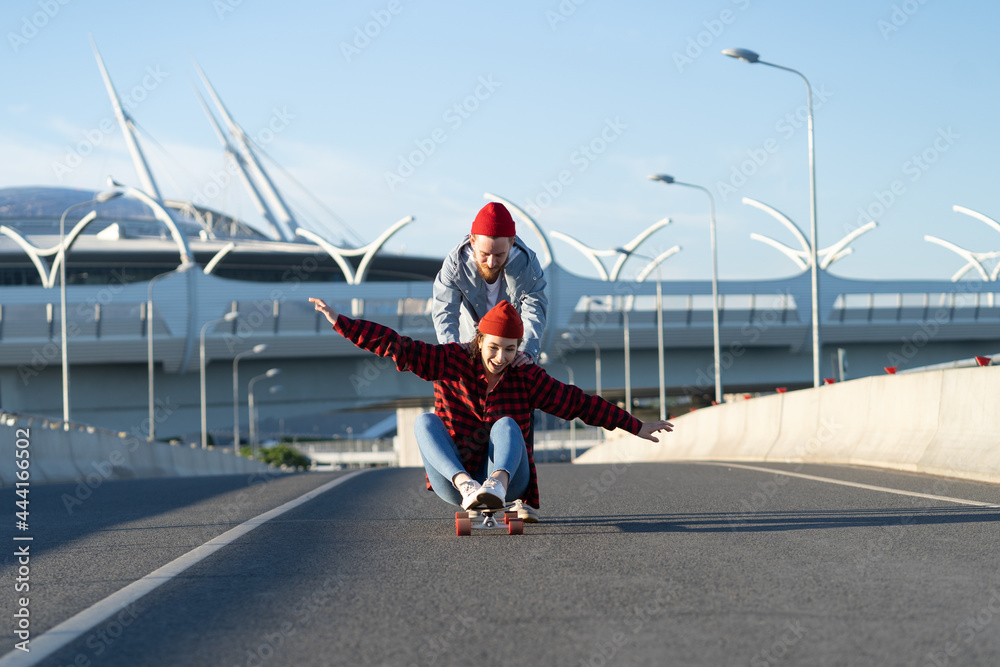Happy carefree woman sit on longboard while man pushing her back riding on empty city street. Young couple of hipsters skateboarders enjoy time outdoors together have fun longboarding. Freedom concept Happy carefree woman sit on longboard while man pushing her back riding on empty city street. Young couple of hipsters skateboarders enjoy time outdoors together have fun longboarding. Freedom concept