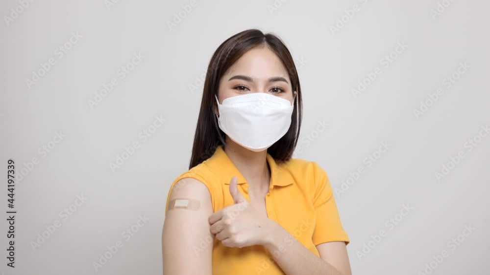Beautiful asian woman in yellow shirt wearing mask getting a vaccine protection the coronavirus. Happy female pointing arm with bandage after receiving vaccination and thumbs up. Beautiful asian woman in yellow shirt wearing mask getting a vaccine protection the coronavirus. Happy female pointing arm with bandage after receiving vaccination and thumbs up.