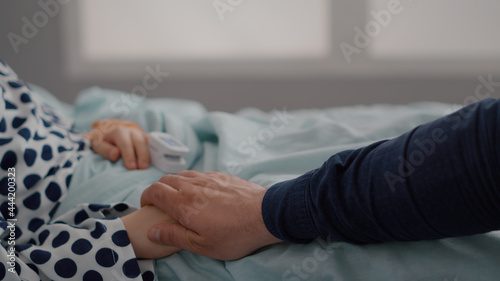 Closeup of sick child resting in bed with oximeter on finger monitoring heartbeat pulse after suffering recovery surgery. Father holding daughter hands during sickness examination in hospital ward