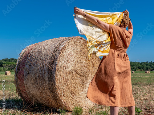 A woman standing in a field with hay bales partially covers her head with a white and yellow scarf photo