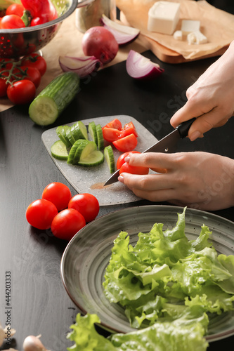 Woman cutting vegetables for tasty Greek salad on dark wooden background