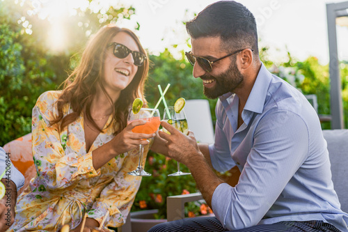 Canvas Print Couple of young friends having a celebratory toast with clinking glasses with fruit cocktails while sitting on a sofa at a garden party