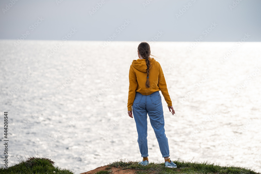 A woman traveler in a bright yellow sweatshirt and loose jeans stands on the seashore and looks into the distance. Freedom and a sense of tranquility. Love of travel