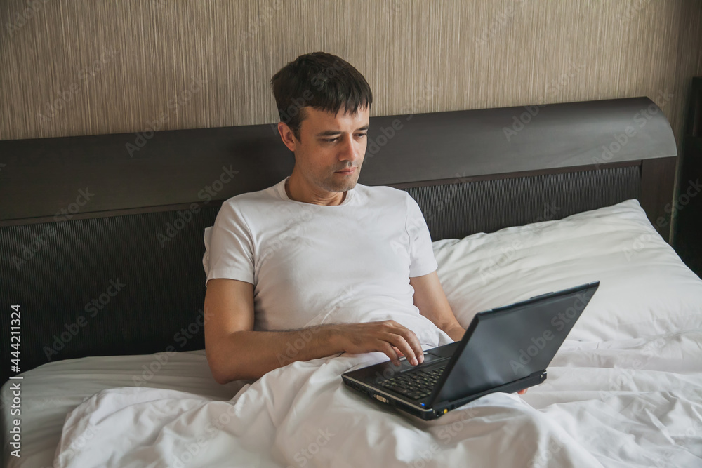 A middle-aged man is sitting in bed in his bedroom and working on a laptop A middle-aged man is sitting in bed in his bedroom and working on a laptop