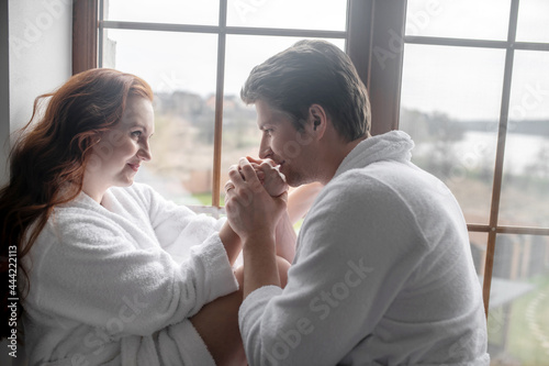 A couple in white bathrobes standing near the window and looking romantic