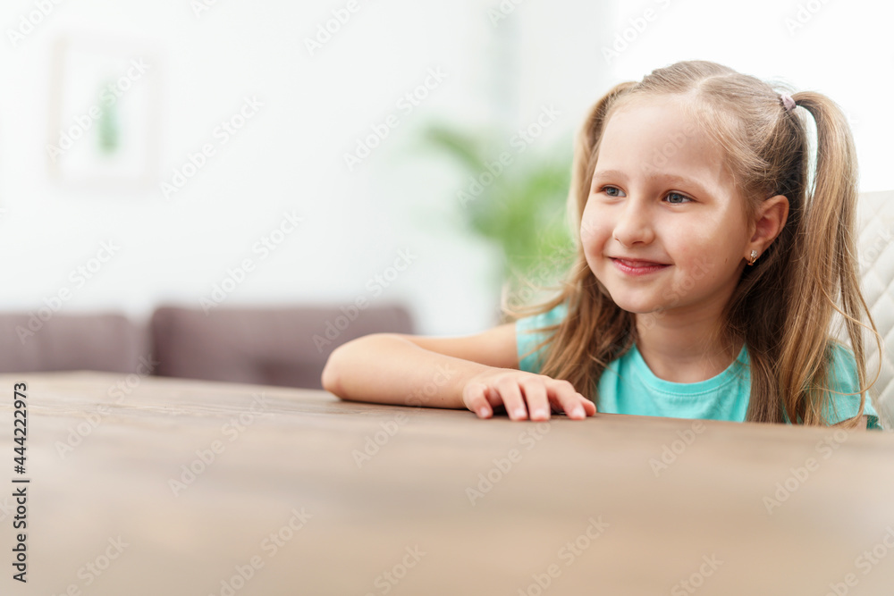 little girl with brown hair is sitting at home at the table and smiling cheerfully against the background of a bright room. little girl with brown hair is sitting at home at the table and smiling cheerfully against the background of a bright room.