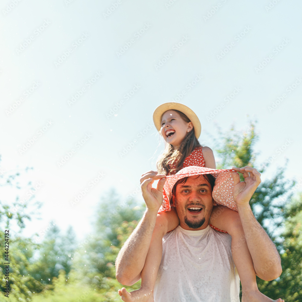 happy little preschool girl is having fun, laughing, riding on her dad's shoulders in the backyard garden on a warm sunny day. Active family games in the fresh air with water splashes. Copy space