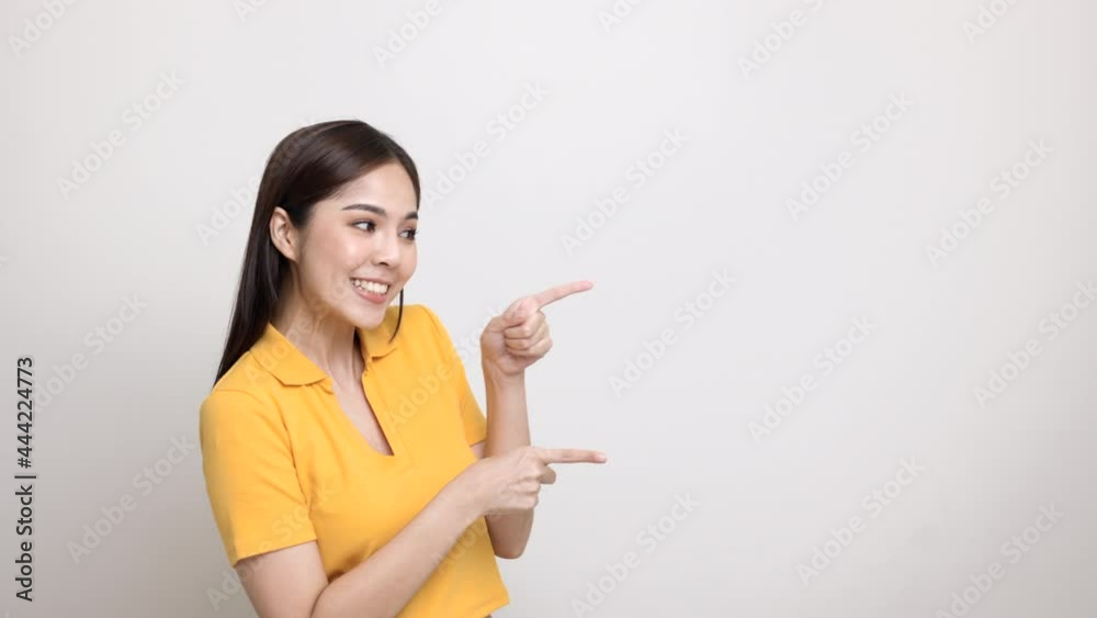 Excited asian woman pointing the finger to blank space for advertising text on isolated background. Joyful teenage girl in yellow shirt standing in white room looking at empty space. Excited asian woman pointing the finger to blank space for advertising text on isolated background. Joyful teenage girl in yellow shirt standing in white room looking at empty space.