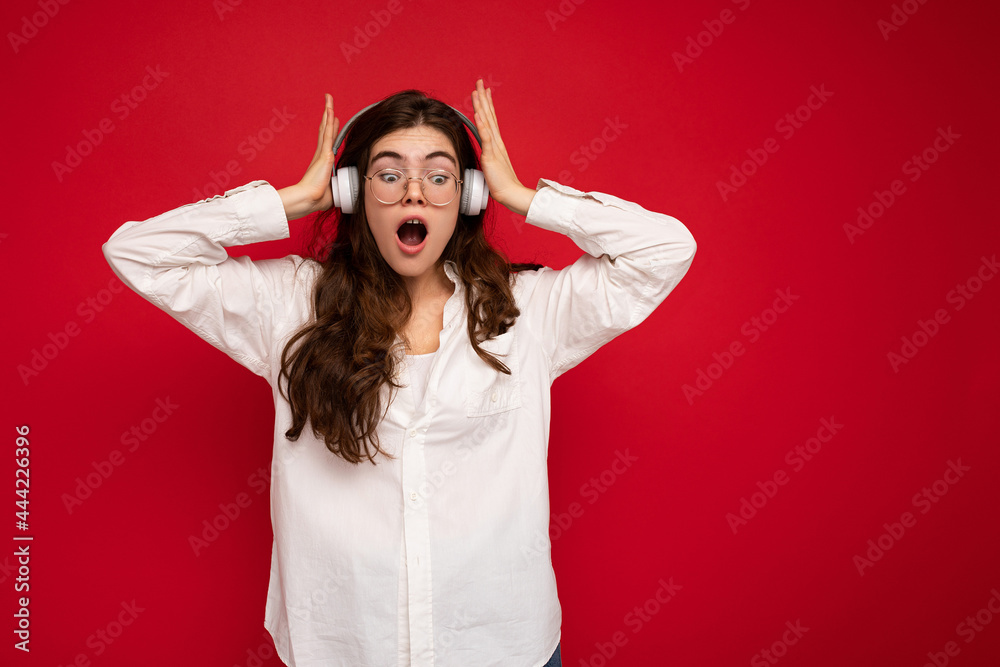 Attractive shocked young brunette female person wearing white shirt and optical glasses isolated over red background wearing white wireless bluetooth earphones listening to music and looking down to Attractive shocked young brunette female person wearing white shirt and optical glasses isolated over red background wearing white wireless bluetooth earphones listening to music and looking down to