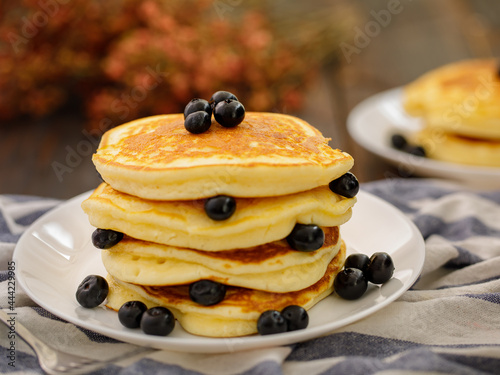 Stack of delicious pancakes sweet food with blueberries and blur dry leaves, plaid fabric in white plate on wooden background.