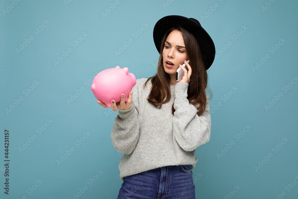 Photo of asking serious thoughtful young attractive pretty woman with dark streight long hair wearing casual grey sweater and black hat isolated over blue background with free space, holds pink pig Photo of asking serious thoughtful young attractive pretty woman with dark streight long hair wearing casual grey sweater and black hat isolated over blue background with free space, holds pink pig