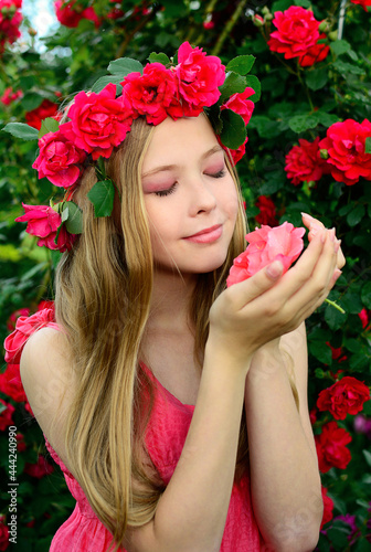 A young girl in a pink dress with a wreath of roses on her head holds a flower with closed eyes in her hands. Vertical
