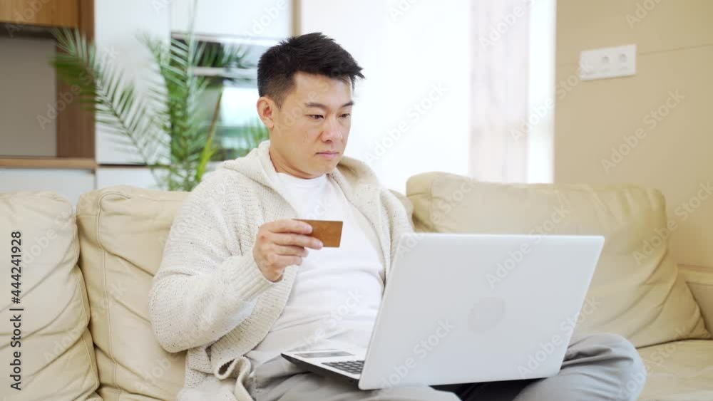 young happy asian man shopping online in internet store using laptop computer and credit card. male enters data of paying for services or tickets in application sitting on couch at home living room young happy asian man shopping online in internet store using laptop computer and credit card. male enters data of paying for services or tickets in application sitting on couch at home living room