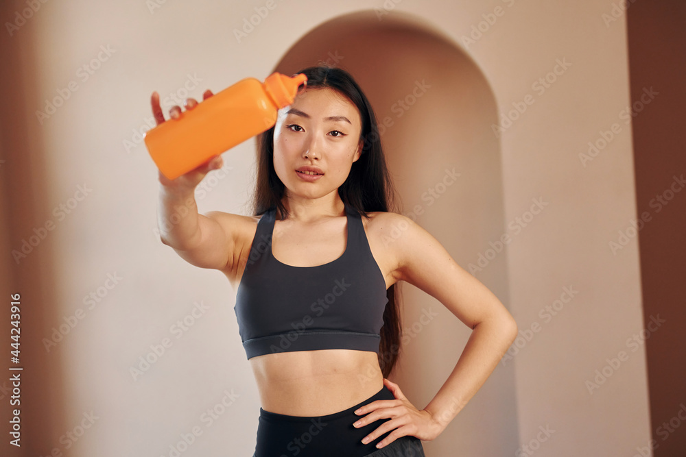 Bottle of water. Young serious asian woman standing indoors Bottle of water. Young serious asian woman standing indoors
