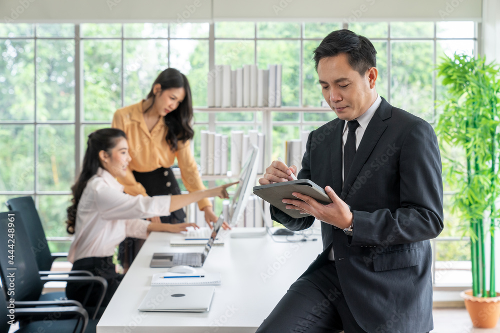Portrait of happy young businessman looking at business document in tablet while his colleagues working in office background on business day. Digital tablet communication and Coworking Space concepts. Portrait of happy young businessman looking at business document in tablet while his colleagues working in office background on business day. Digital tablet communication and Coworking Space concepts.
