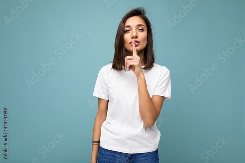 Portrait of young attractive brunette woman with sincere emotions wearing casual white t-shirt for mockup isolated on blue background with copy space and showing shh gesture. Keep secret photo