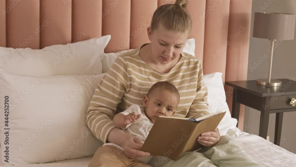 Medium long of blonde Caucasian woman sitting in bed, reading book to her Mixed-Race baby at daytime Medium long of blonde Caucasian woman sitting in bed, reading book to her Mixed-Race baby at daytime