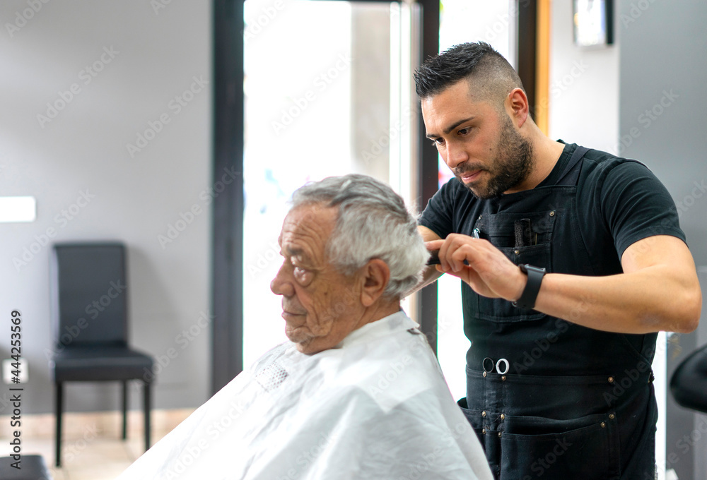 Fototapeta premium Young barber cutting old man's hair with scissors having a good time. Close up