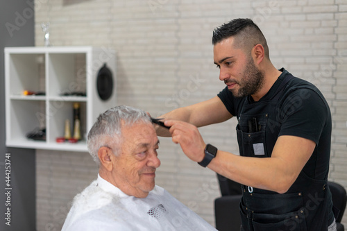 Young barber cutting hair with scissors and comb to an old man in a barber shop.