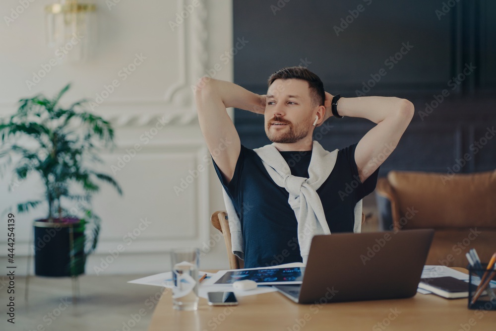 Experienced European man rests on chair looks away with thoughtful expression poses at desktop poses at home office listens information via earphones uses free internet connection prepares for exam