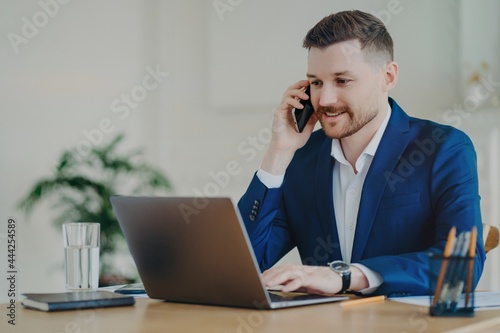 Smiling young attractive businessman talking on phone in front of laptop