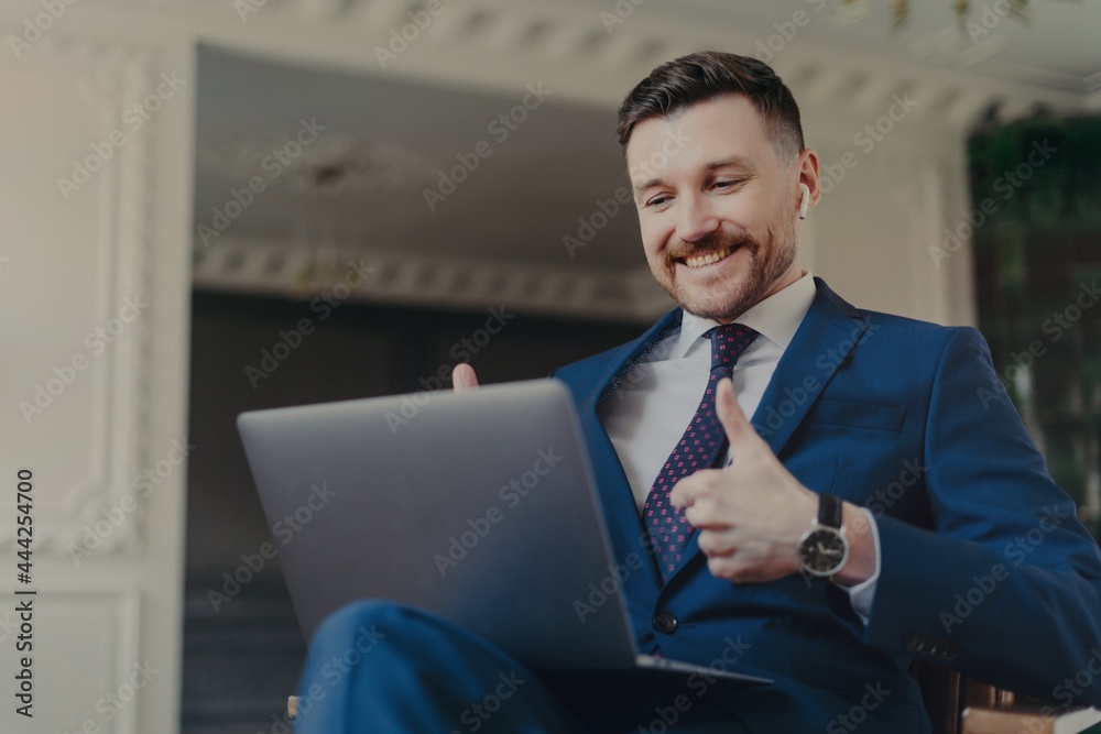 Successful bearded male entrepreneur dressed in formal suit uses modern laptop computer and earbuds for making web conference chatting keeps thumbs raised makes like gesture wears blue formal suit Successful bearded male entrepreneur dressed in formal suit uses modern laptop computer and earbuds for making web conference chatting keeps thumbs raised makes like gesture wears blue formal suit