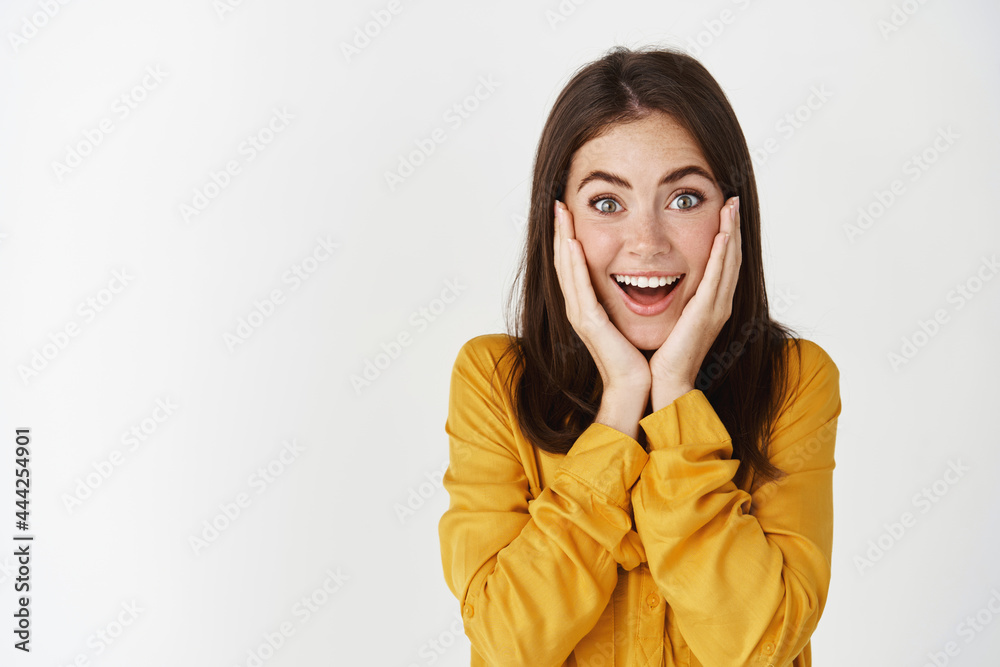 Close-up of happy and surprised woman touching face, staring at camera amazed, standing over white background Close-up of happy and surprised woman touching face, staring at camera amazed, standing over white background