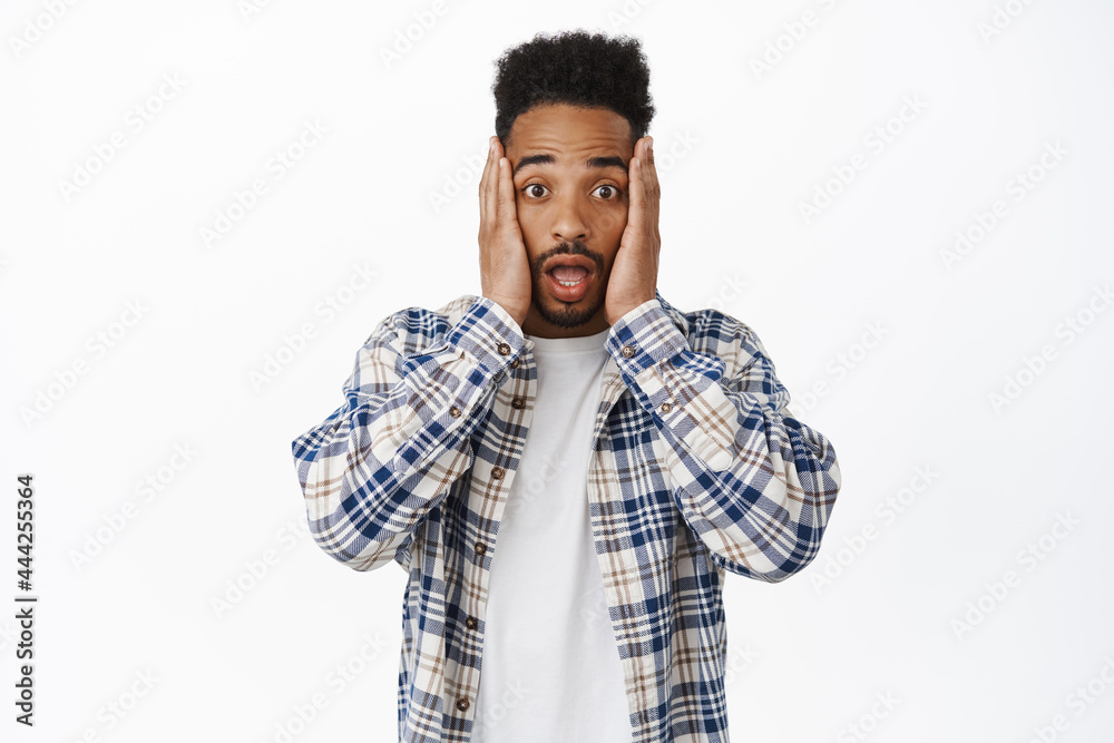 Shocked and worried african american man holding hands on cheeks face, gasp and stare nervously at camera, hear shocking news, standing over white background Shocked and worried african american man holding hands on cheeks face, gasp and stare nervously at camera, hear shocking news, standing over white background