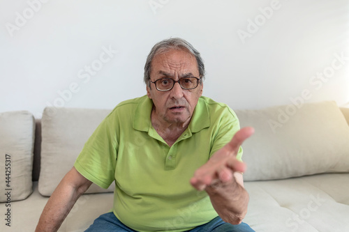 Shot of a senior Caucasian man looking sad and grumpy in his home during the day. Angry elderly man with gray hair wearing eyeglasses looking at camera, sitting on sofa at home. Communication concept.