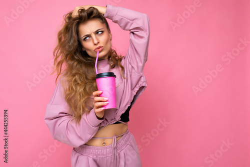 Photo shot of beautiful sad upset young blonde woman wearing pink sport clothes isolated over pink background holding paper coffee cup for mockup drinking and looking to the side