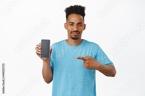 Disappointed african american guy pointing finger at smartphone screen with dislike, furrow eyebrows displeased, dislike interface, bad application, standing in t-shirt against white background