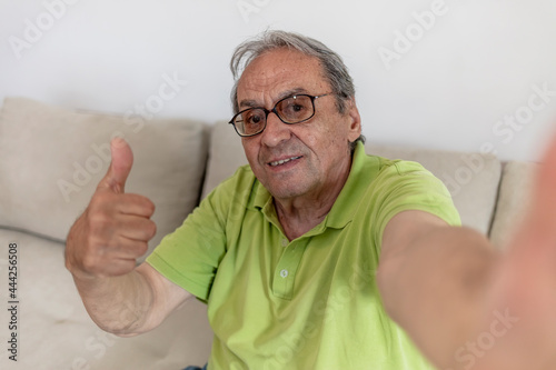 Grandfather taking selfies at home in the living room. Close up portrait of happy cheerful delightful elderly man taking a selfie. Retirement and people concept. Old man taking selfie inside his home.
