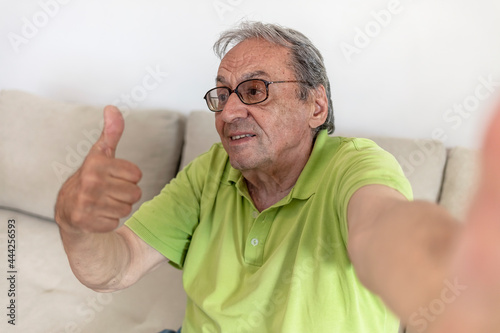 Senior man taking selfie in the comfort of his home. Close up of a man using phone in the living room. Selfie portrait of happy old man with gray hair. Elderly man photographed his face, toothy smile.