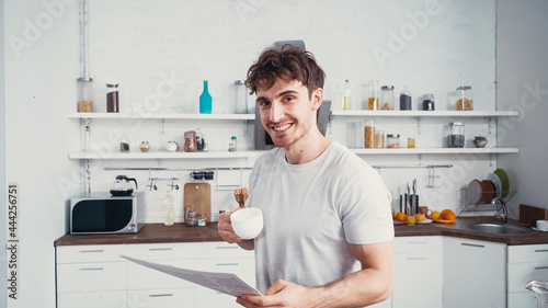happy man with cup of coffee and morning newspaper smiling at camera in kitchen