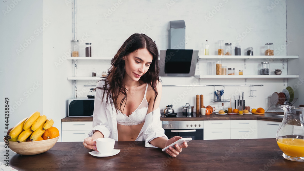 sexy woman looking at smartphone near cup of coffee, fresh fruits and orange juice