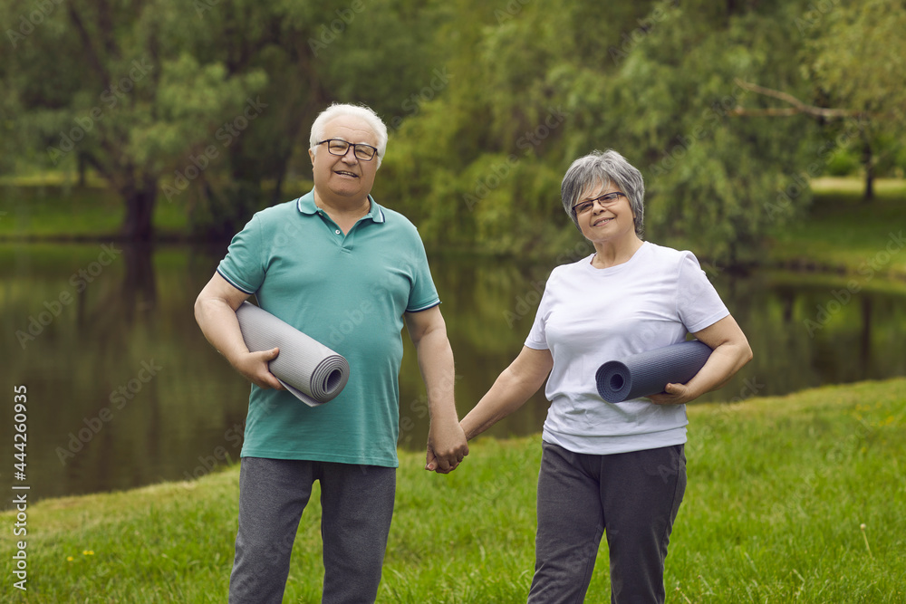 Portrait of happy senior couple in sports clothes with exercise rubber mats holding hands, smiling and looking at camera standing on green lawn after fitness workout and yoga practice in nature Portrait of happy senior couple in sports clothes with exercise rubber mats holding hands, smiling and looking at camera standing on green lawn after fitness workout and yoga practice in nature