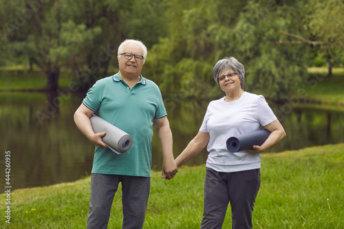 Portrait of happy senior couple in sports clothes with exercise rubber mats holding hands, smiling and looking at camera standing on green lawn after fitness workout and yoga practice in nature