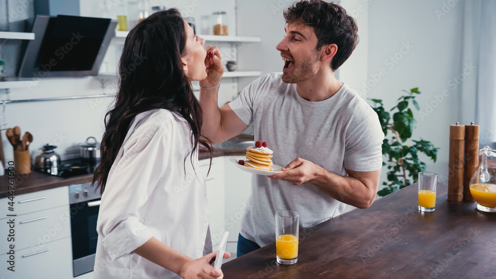 Fototapeta premium cheerful man holding pancakes and feeding girlfriend with raspberry in kitchen
