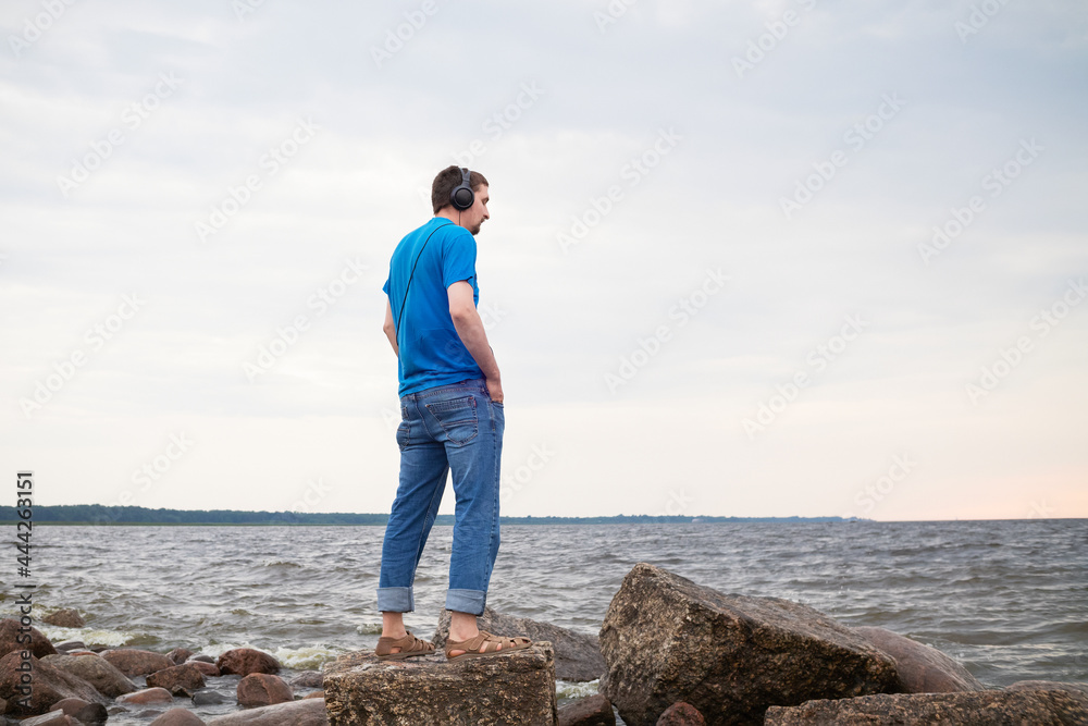 Man with headphones standing in profile on rocky seashore in blue t-shirt and jeans.