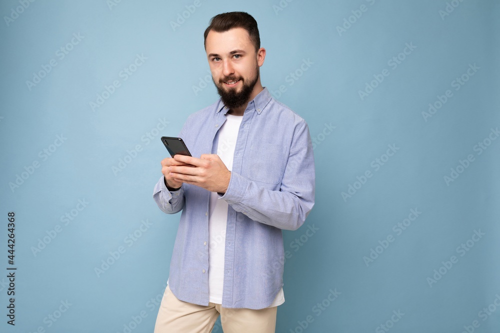 handsome young brunette unshaven man with beard wearing stylish white t-shirt and blue shirt isolated over blue background with empty space holding in hand and using phone messaging sms looking at handsome young brunette unshaven man with beard wearing stylish white t-shirt and blue shirt isolated over blue background with empty space holding in hand and using phone messaging sms looking at